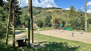 Um parquinho com balanços e um escorregador, rodeado por gramado e árvores, com uma paisagem montanhosa ao fundo.