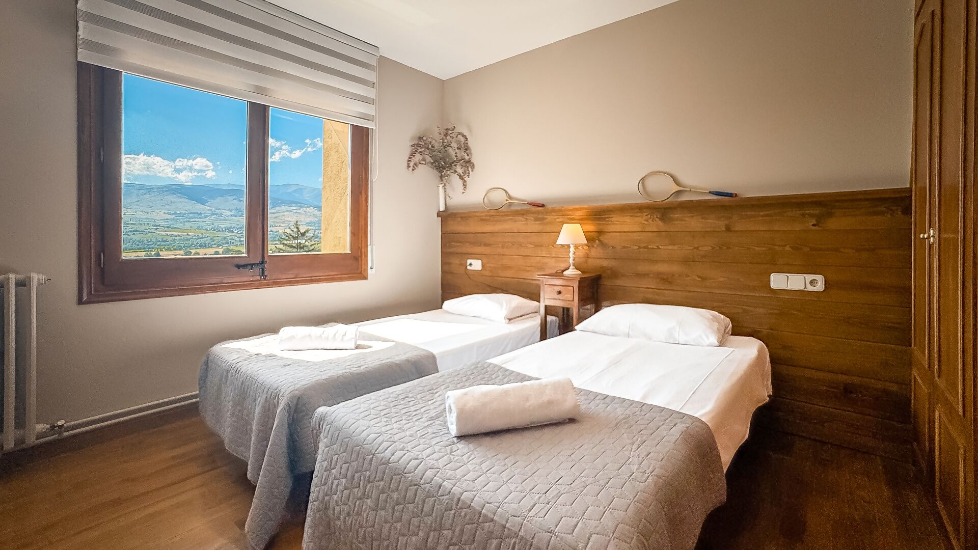 A room with two beds, a wooden headboard, a window with a view, and a nightstand with a lamp.