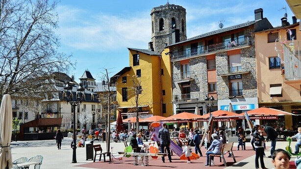 A bustling square with a yellow building, a stone tower, and outdoor seating areas. - Cal Gatesques 1 (Puigcerda)