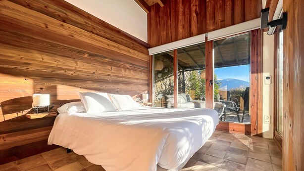 Une chambre avec un plafond en bois, un lit recouvert de draps blancs, une table de chevet avec une lampe, et une vue sur l’extérieur grâce à une porte-fenêtre coulissante.