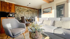 Un salon moderne avec un mur en pierre, un canapé gris, un fauteuil gris et un coin repas avec une table dressée pour quatre personnes.