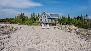A large house with a deck and a covered area, surrounded by a rocky beach and trees.