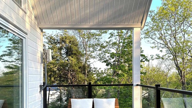 A covered outdoor seating area with a wooden deck, a cushioned bench, and two white lounge chairs.