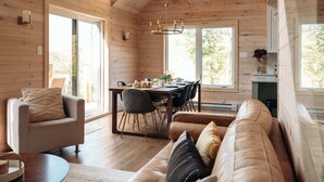 A cozy cabin interior with a dining area, a sofa, and a large window providing natural light. - Montagnia escape, Tremblant, Private Spa & Sauna Retreat (Val-des-Lacs)