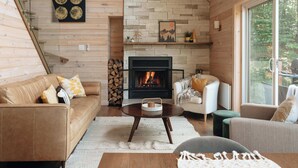 A cozy cabin interior with a fireplace, wooden walls, and a large window offering a view of the outdoors. - Montagnia escape, Tremblant, Private Spa & Sauna Retreat (Val-des-Lacs)