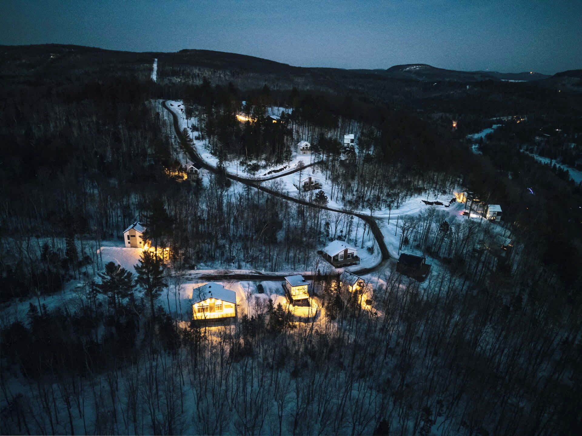 An aerial view of a snow-covered village with several houses and a church, surrounded by forested hills.