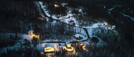 An aerial view of a snow-covered village with several houses and a church, surrounded by forested hills.
