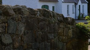 Un bâtiment blanc avec un toit à forte pente, un mur de pierre au premier plan et des arbres en arrière-plan.