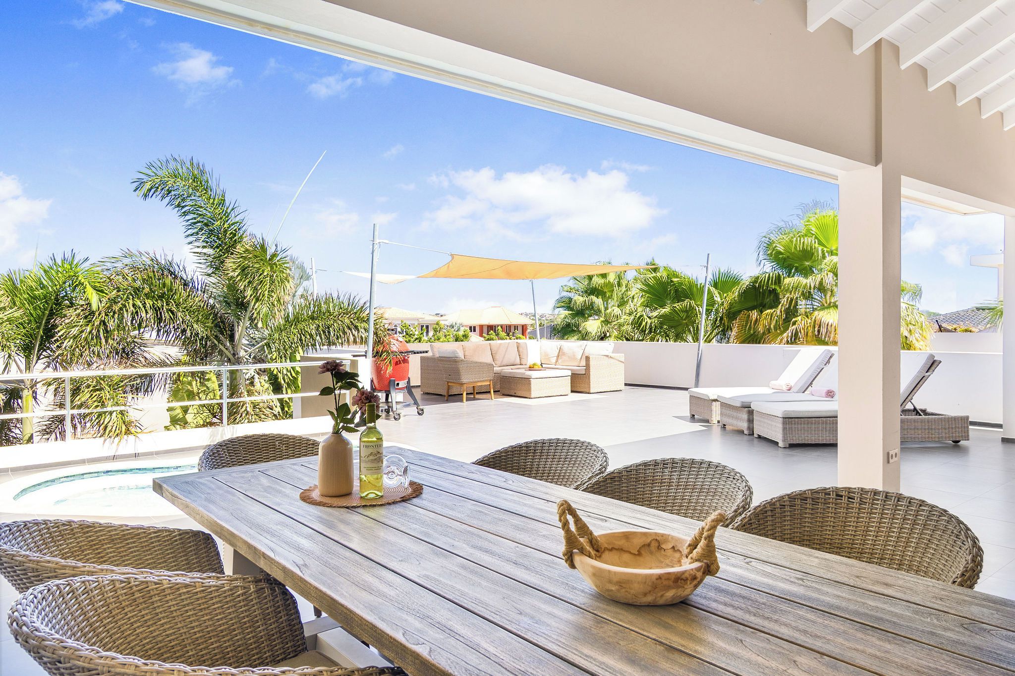 An outdoor patio with a wooden table, wicker chairs, a lounge area with cushions, and a view of palm trees and a swimming pool.