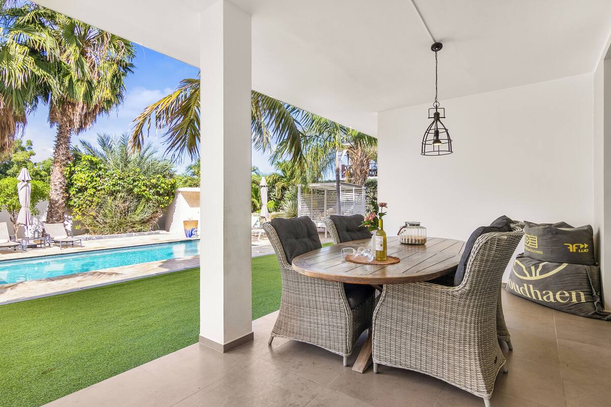 A patio with a dining table and chairs, a hanging light fixture, and a view of a swimming pool and greenery.