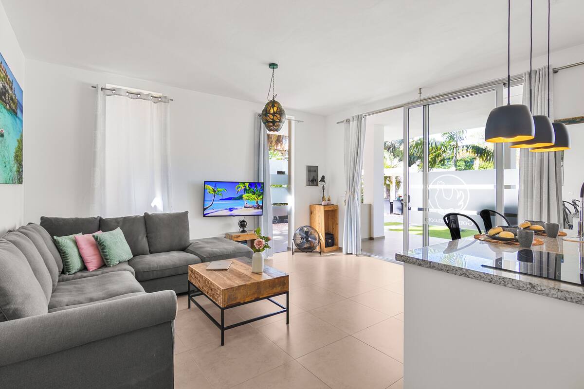 A modern living room with a grey sofa, a wooden coffee table, a flat-screen TV, and a large window with a view of greenery.
