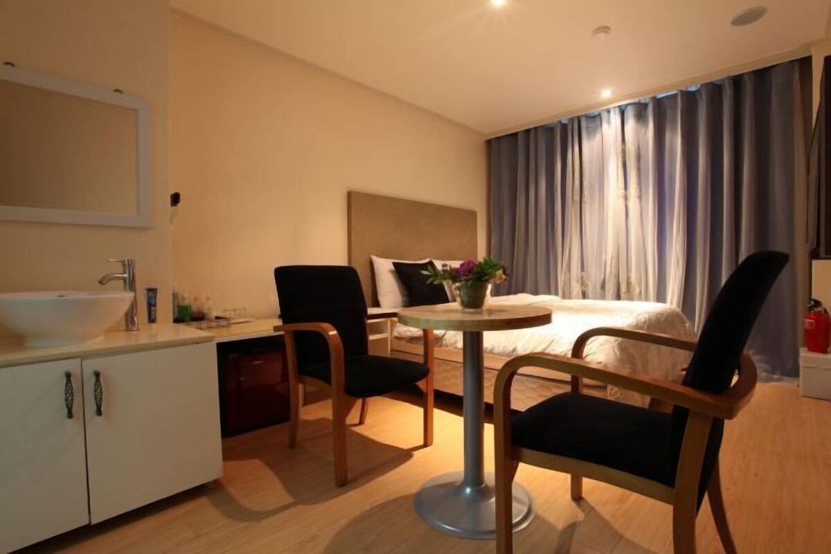A hotel room with a bed, a bedside table with a lamp, a chair, a small table with a vase, and a bathroom with a sink and cabinet.