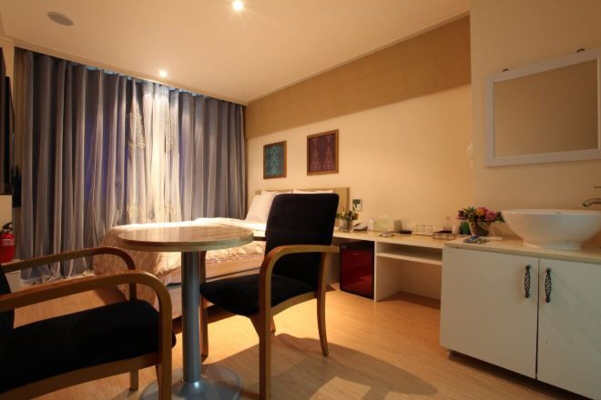 Habitación de hotel con cama, una mesa redonda, una silla, un escritorio, un mueble blanco con lavabo y una ventana con cortinas.