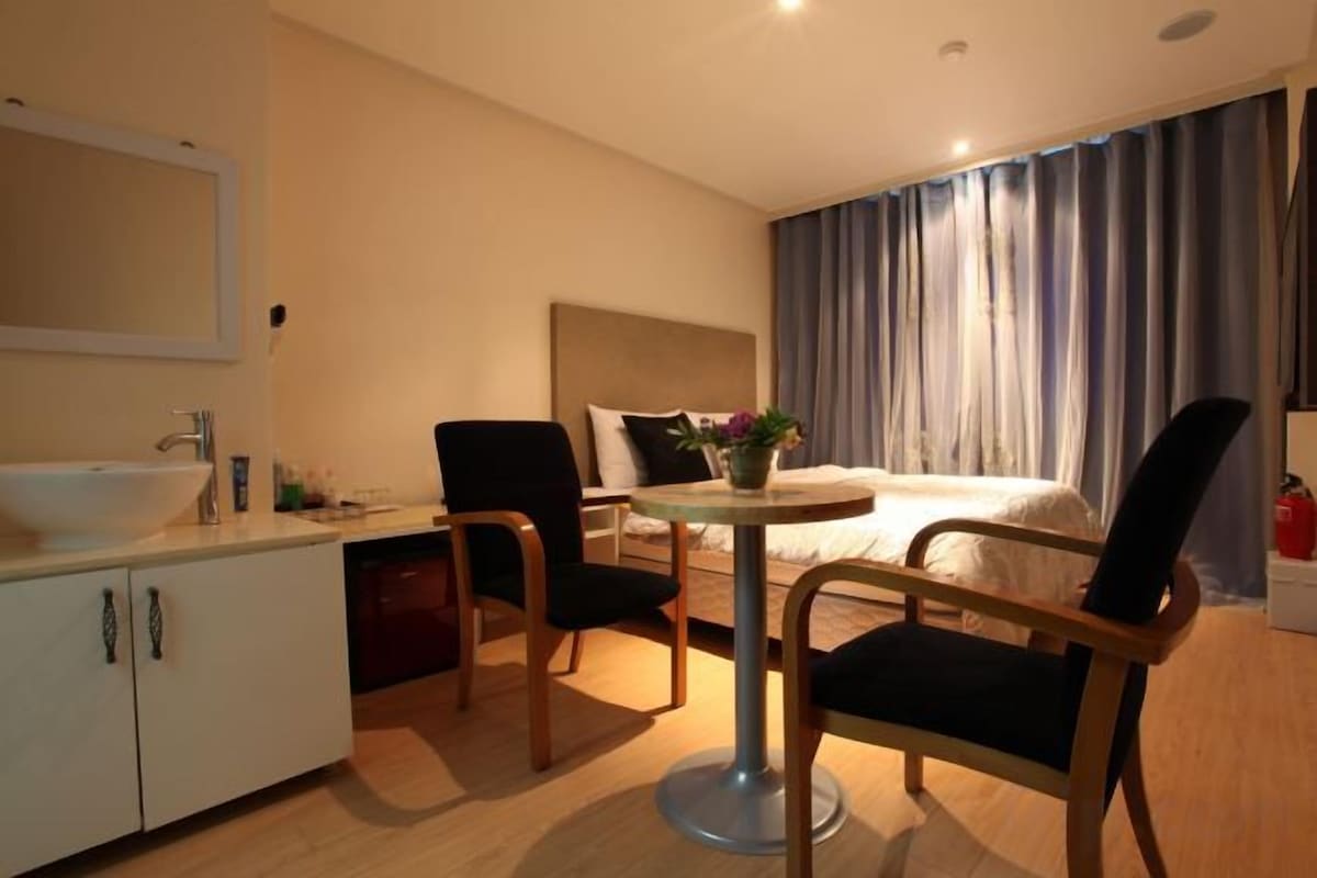 Une chambre d’hôtel avec un lit, un bureau, une chaise, un lavabo et une plante en pot.