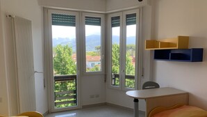 Ein Zimmer mit zwei Betten, einem Schreibtisch und Blick auf Grünflächen und Gebäude durch große Fenster.