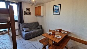Une pièce de taille modeste, comprenant un canapé, une table en bois avec de l’assiette et des couverts, un luminaire suspendu et une fenêtre avec des rideaux.