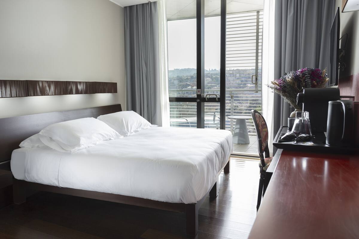 Ein Hotelzimmer mit einem großen Bett, einem Schreibtisch mit Kaffeemaschine, einem Sessel und Blick auf die Stadt durch große Fenster.