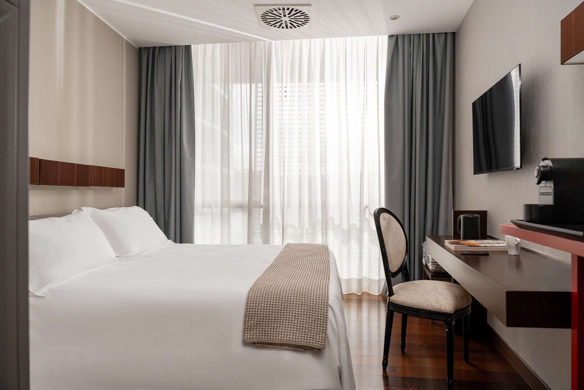 A hotel room with a bed, a desk with a chair, a TV, and a window with curtains.