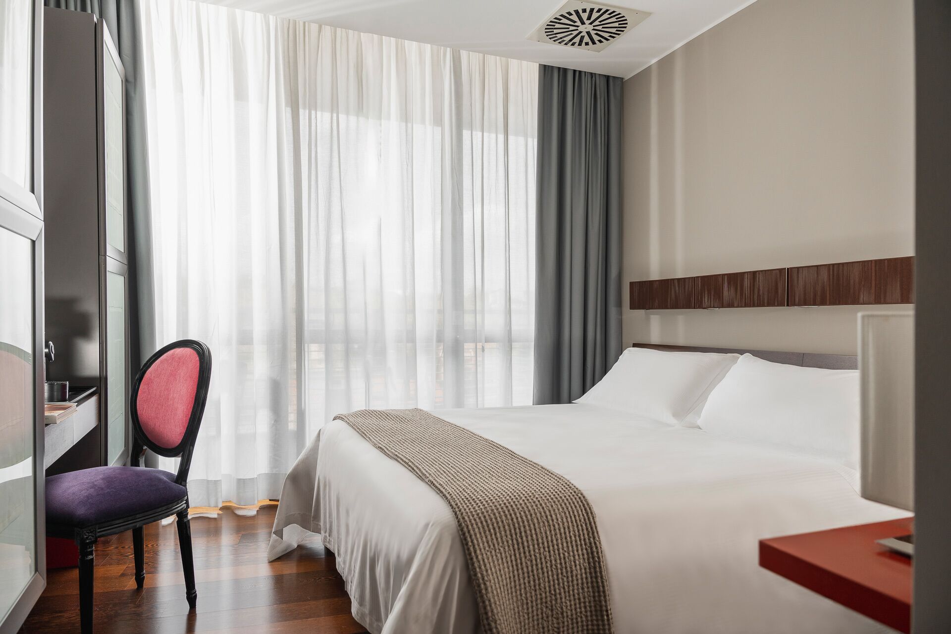 Ett hotellrum med en säng, ett skrivbord med en stol och ett stort fönster med gardiner.