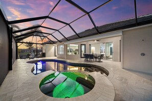Zona de piscina moderna al aire libre con techo de cristal, rodeada por un patio con asientos y vistas al atardecer.