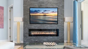 Ein modernes Wohnzimmer mit Kamin, einem flachbildfernseher darüber und einem Glastisch im Vordergrund.