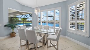 Ein moderner Essbereich mit einem Glastisch und weißen Stühlen, großen Fenstern mit Fensterläden und Blick auf einen Swimmingpool.