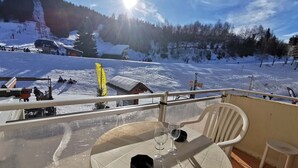 A balcony with a table and chairs overlooking a snowy mountain area with ski slopes and a building in the background. - Charming Apartment Near Ski Lifts in Prapoutel, Sleeps 6, Balcony, TV, No Pets (Les Adrets-Prapoutel)