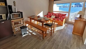 A cozy living room with a wooden dining table, a sofa with red cushions, a clock on the wall, and a view of the sea through large windows. - Charming Apartment Near Ski Lifts in Prapoutel, Sleeps 6, Balcony, TV, No Pets (Les Adrets-Prapoutel)