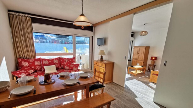 A cozy living area with a dining table set for four, a sofa with red cushions, a television, and a view of a snowy landscape through the window. - Charming Apartment Near Ski Lifts in Prapoutel, Sleeps 6, Balcony, TV, No Pets (Les Adrets-Prapoutel)