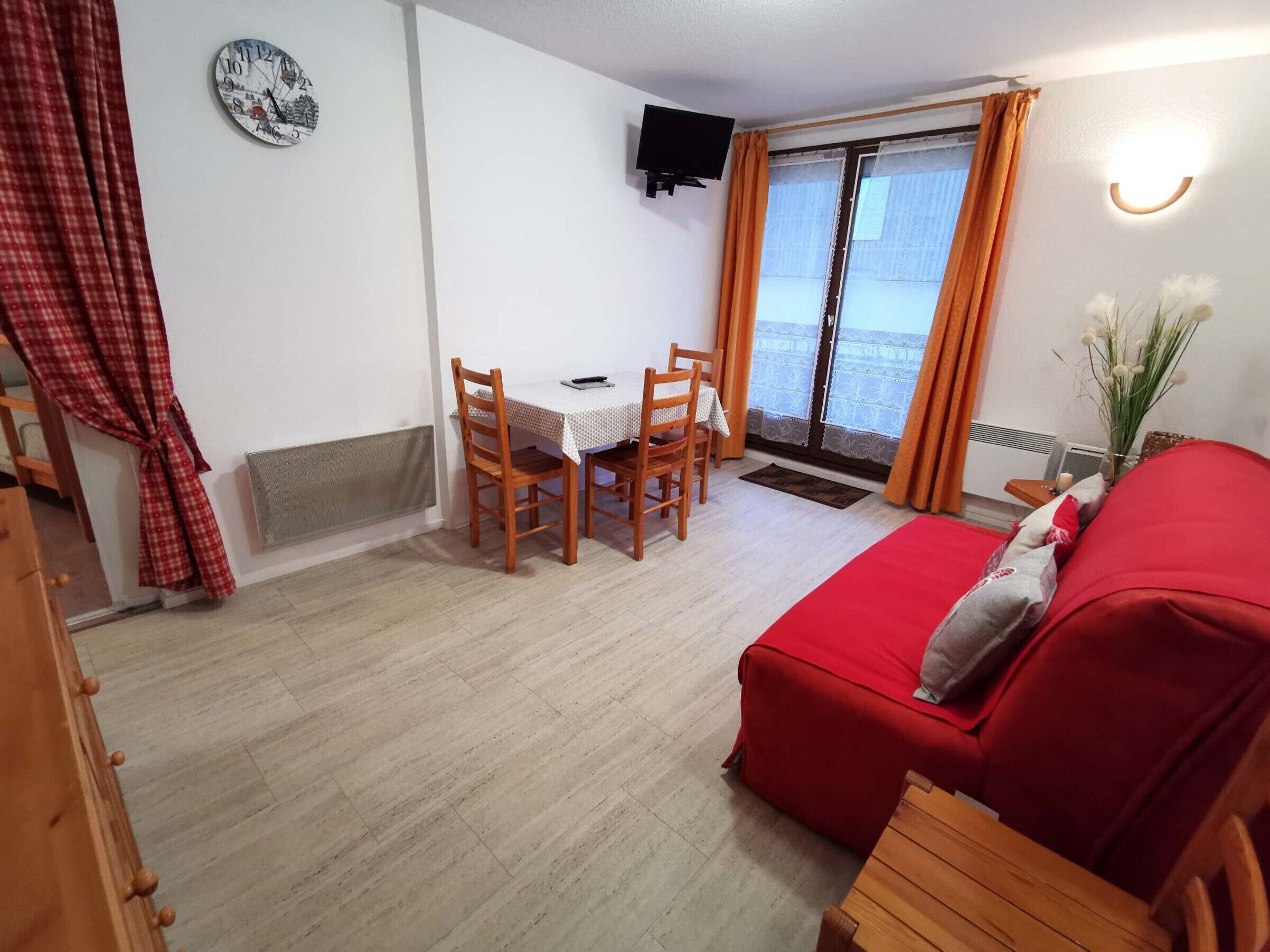 Een kamer met een rode bank, een houten tafel met stoelen, een klok aan de muur en een tv die boven de tafel is bevestigd.