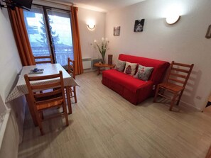 A living room with a red sofa, wooden chairs, a dining table with chairs, a small side table, and a window with curtains.