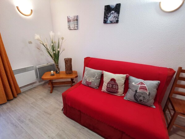 A red sofa with three decorative pillows, a small wooden side table with a candle and a vase of white flowers, and a wall-mounted picture of a mountain landscape.