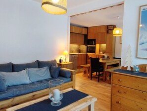 Ein gemütliches Wohnzimmer mit einer Couch, einem Couchtisch und einem Essbereich mit Tisch und Stühlen. Im Hintergrund ist eine Küche mit Holzschränken und Geräten zu sehen.