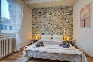 A bedroom with a stone wall, a bed with white bedding, two bedside tables with candles, and a window with white curtains. - La Valbinella Few Meters From Varese Lake (Gavirate)
