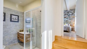 Une salle de bain moderne avec un mur en pierre, une douche en verre et un plancher en bois menant à une chambre également dotée d’un mur en pierre et d’un lit.