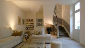 Un salon spacieux avec un canapé blanc, une table basse en verre, une bibliothèque et un escalier menant à un niveau supérieur.