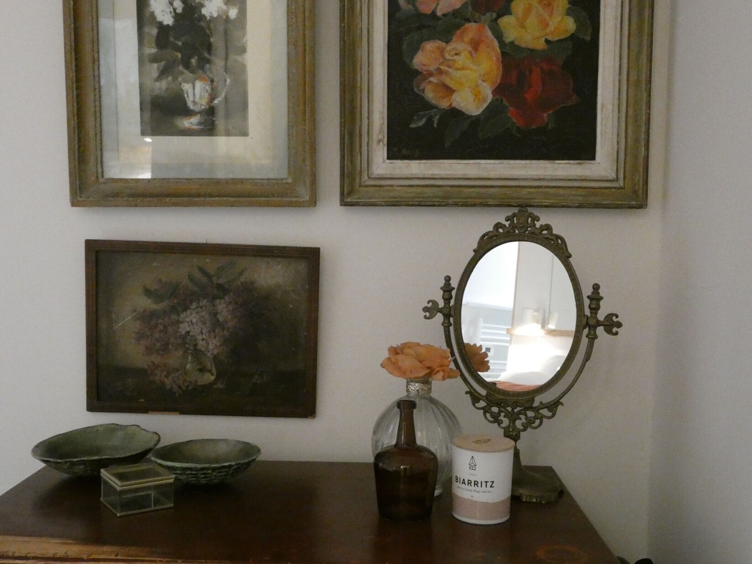 A room with three framed floral paintings on the wall, a vintage mirror, and a table with decorative items including a candle and bowls.