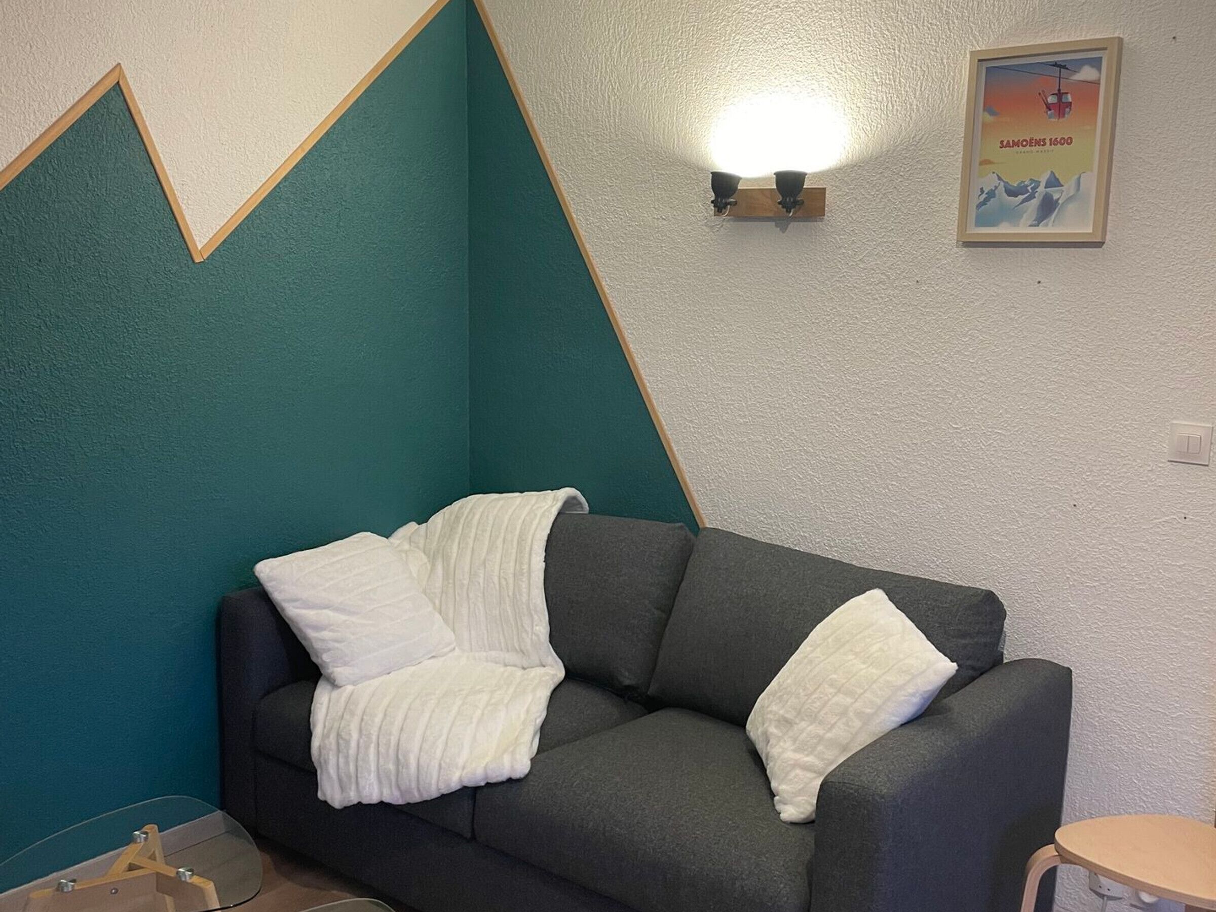 Een kamer met teal wanden, een donkergrijze bank, witte kussens, een wandlamp en een lijst met een afbeelding.