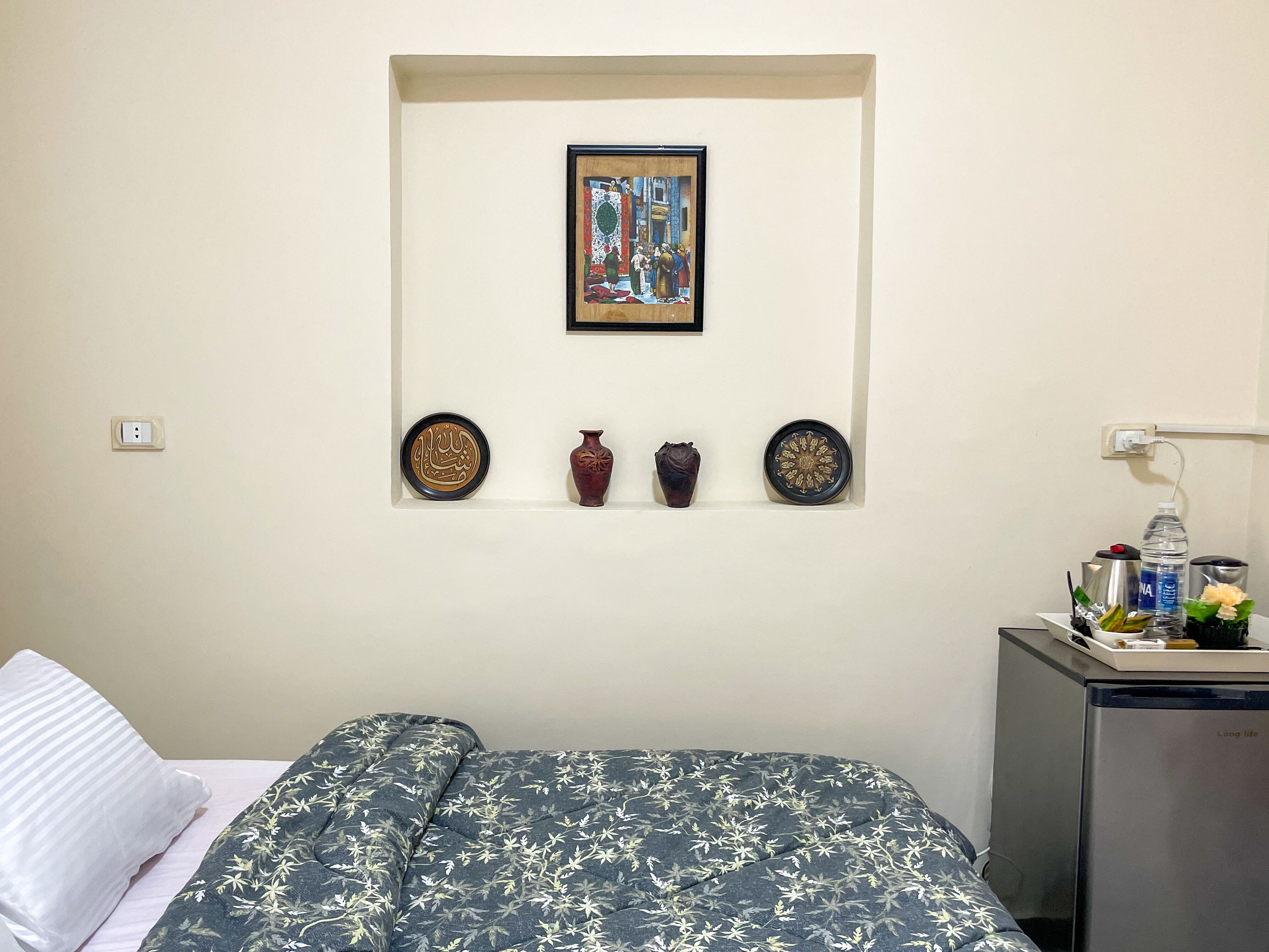 A hotel room with a bed, a small table with a water bottle, and a decorative wall piece above the bed.