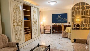 A hotel lobby with a large chandelier, a seating area with sofas, a decorative wall with a sign, and a patterned floor. - Chez l'ami hotel (Giza)