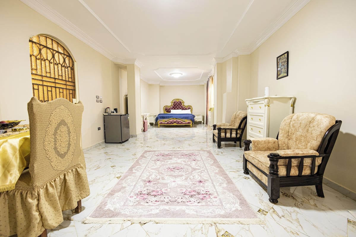 Una habitación amplia con una cama grande, una zona de estar con una silla y una mesa, y una alfombra decorativa.