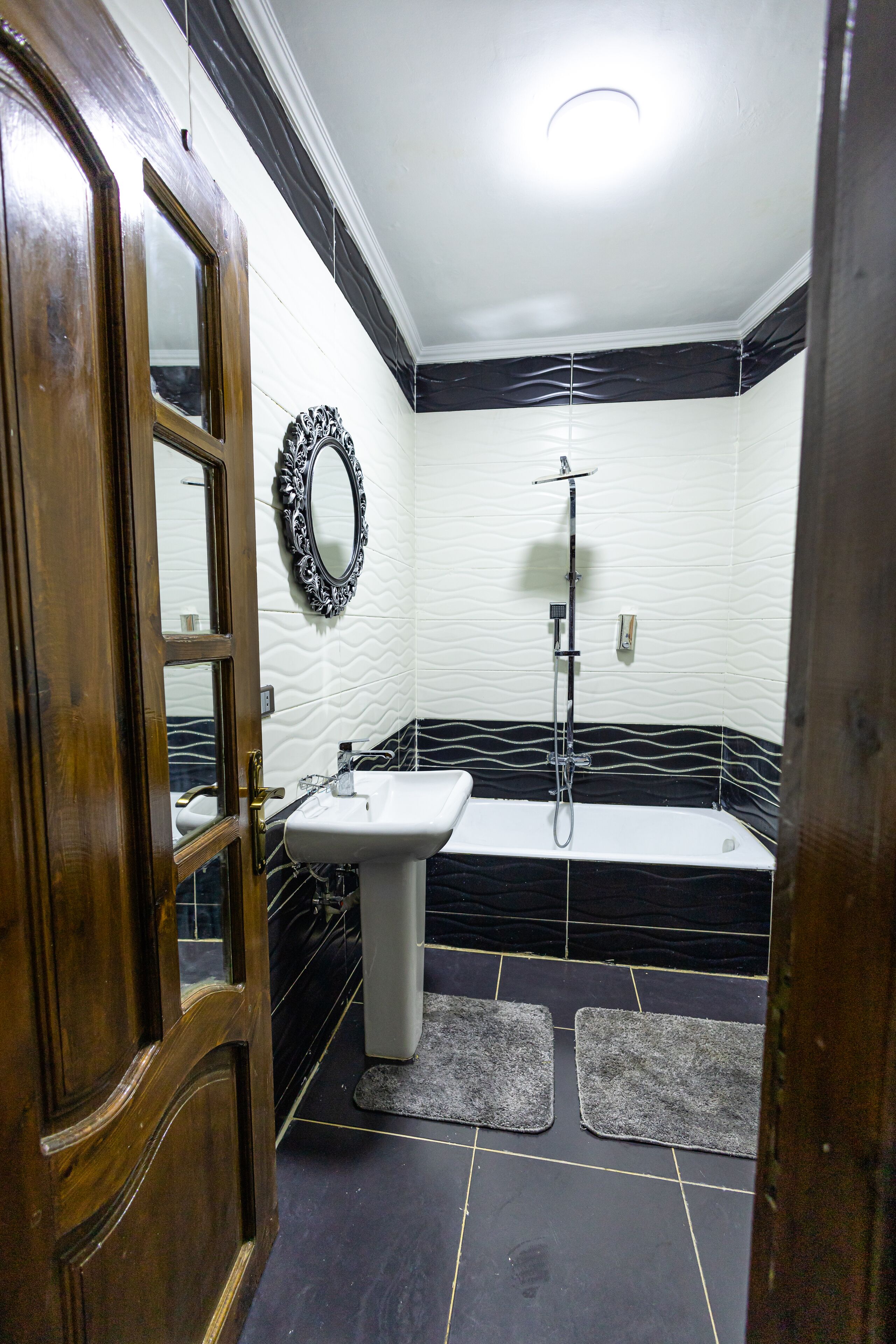 A bathroom with a wooden door, a white sink, a black and white tiled shower, and a mirror on the wall.