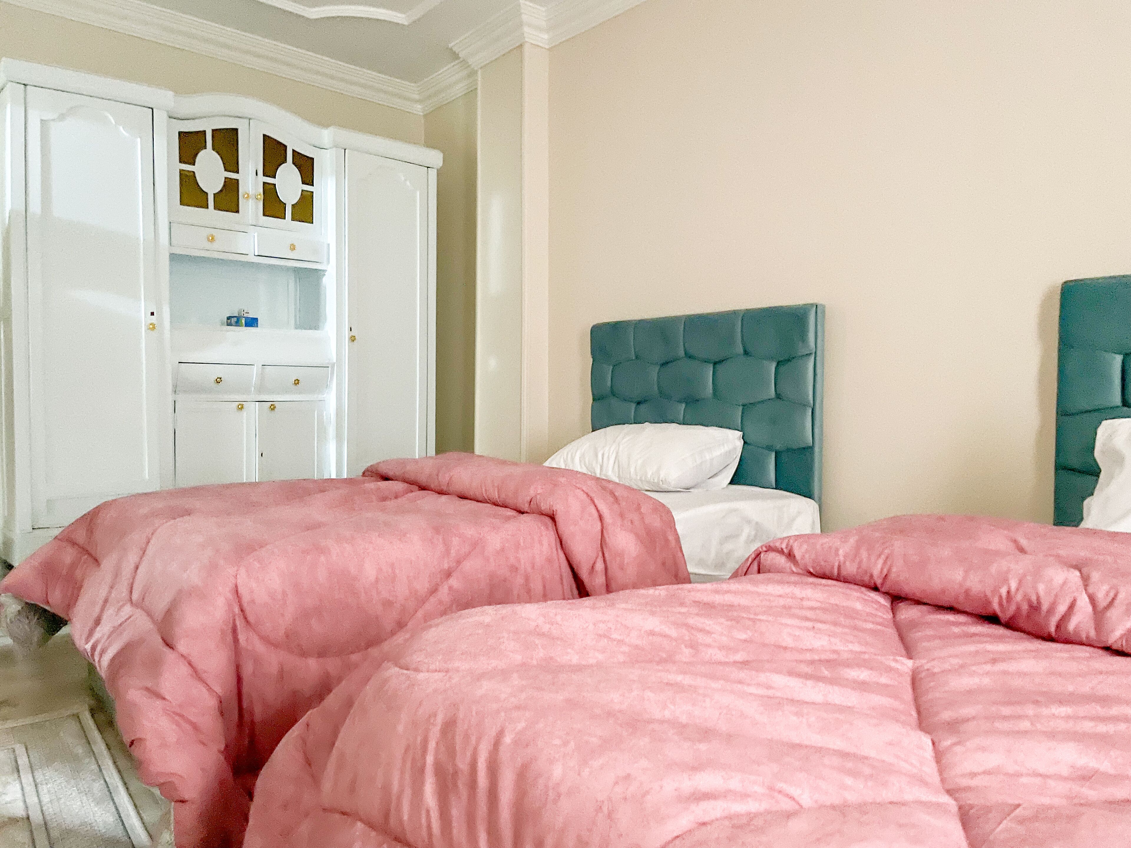 Une chambre avec deux lits, chacun agrémenté de linge de lit rose, d’une tête de lit turquoise et d’un oreiller blanc. On y trouve une armoire blanche avec un panneau décoratif et une petite étagère.