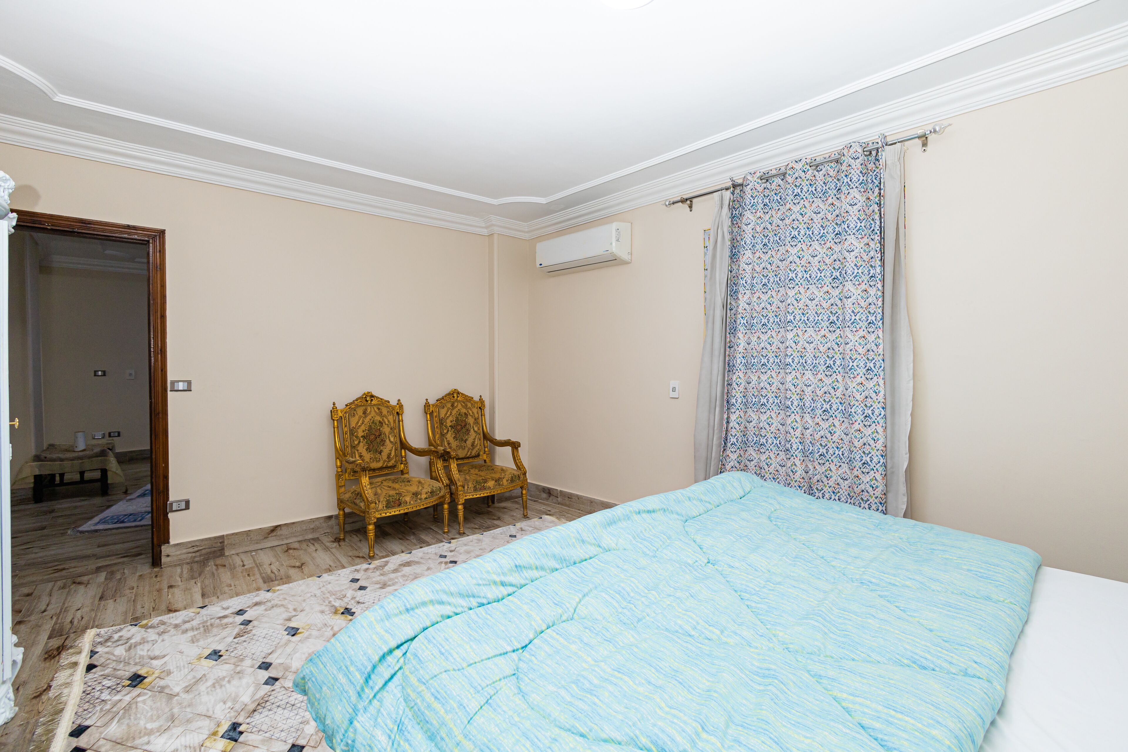 Un dormitorio con una cama, dos sillones decorados, una cortina estampada y un aire acondicionado.