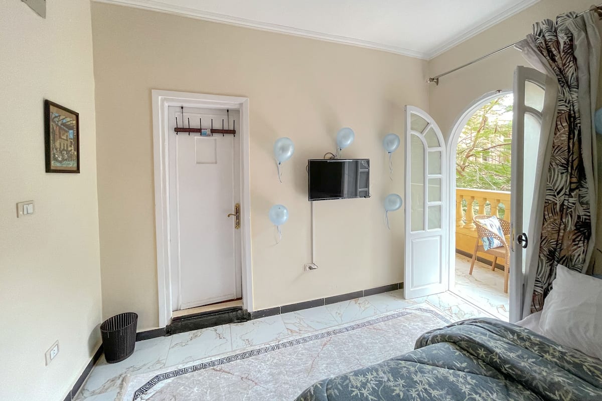 Un dormitorio con cama, puerta, televisor montado en la pared y balcón con una silla.