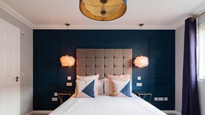 Une chambre à coucher avec un grand lit, une tête de lit capitonnée, deux lampes suspendues et un mur d’accent bleu foncé.