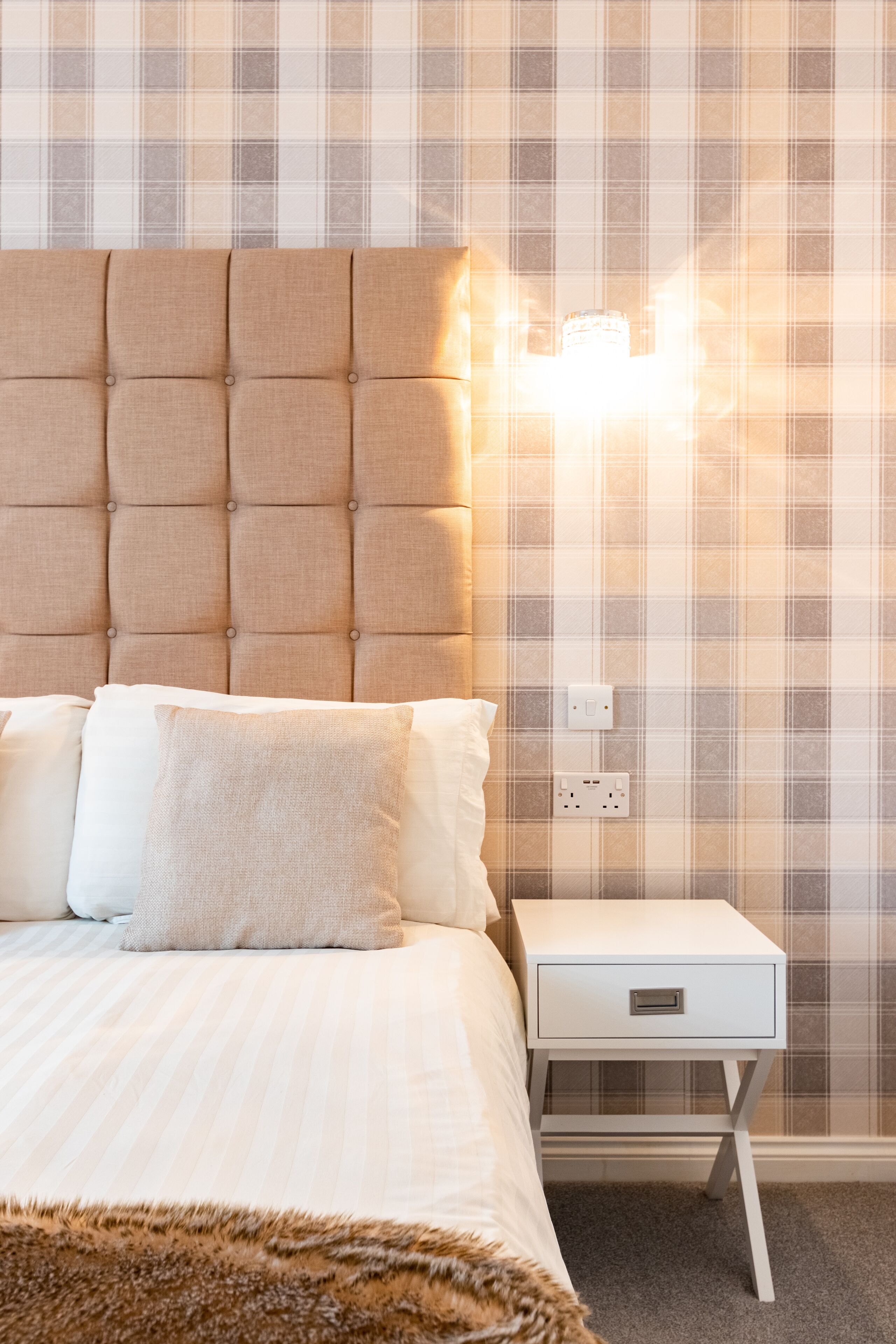 A neatly made bed with a tufted headboard, a bedside table with a drawer, and a wall with a plaid pattern.