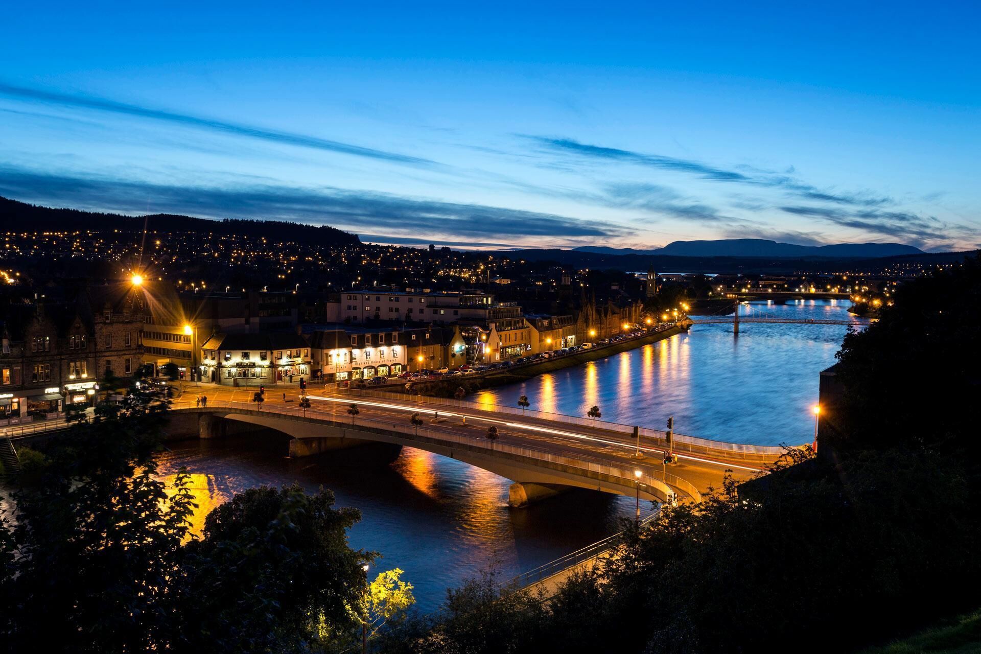 A nighttime view of a riverside town with a bridge, illuminated buildings, and a calm river reflecting lights.