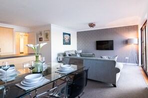 A modern living room with a glass dining table set for a meal, a grey sofa, and a flat-screen TV mounted on the wall.