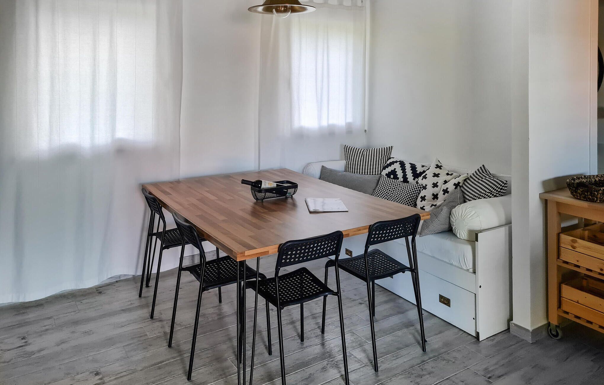 A room with a wooden dining table, four black chairs, a white sofa with patterned cushions, and a small wooden side table.